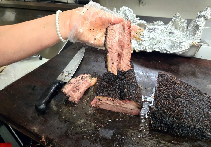 A gloved hand holds up a slice of brisket with a larger brisket on the counter next to a knife.