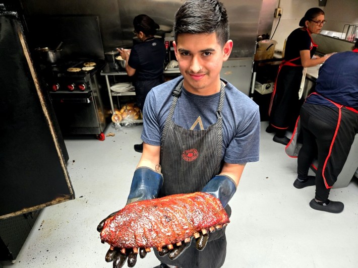 A man with a mustache and a striped apron on holds a rack of ribs.