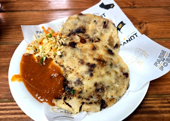 Two black-spotted pupusas with red salsa and curtido on the side, filled with brisket, on a paper plate.