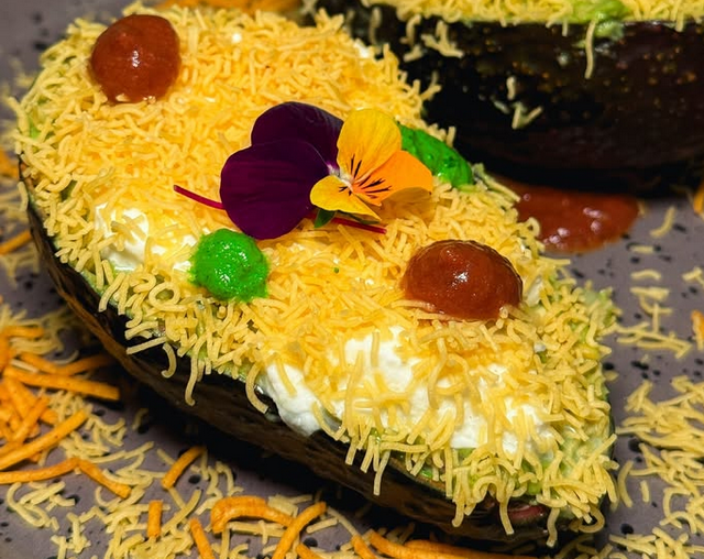 An avocado sliced in half and filled with burrata cheese and tiny, yellow noodes-like nylon sev, with green and orange dots of sauce, and an edible pansy on top